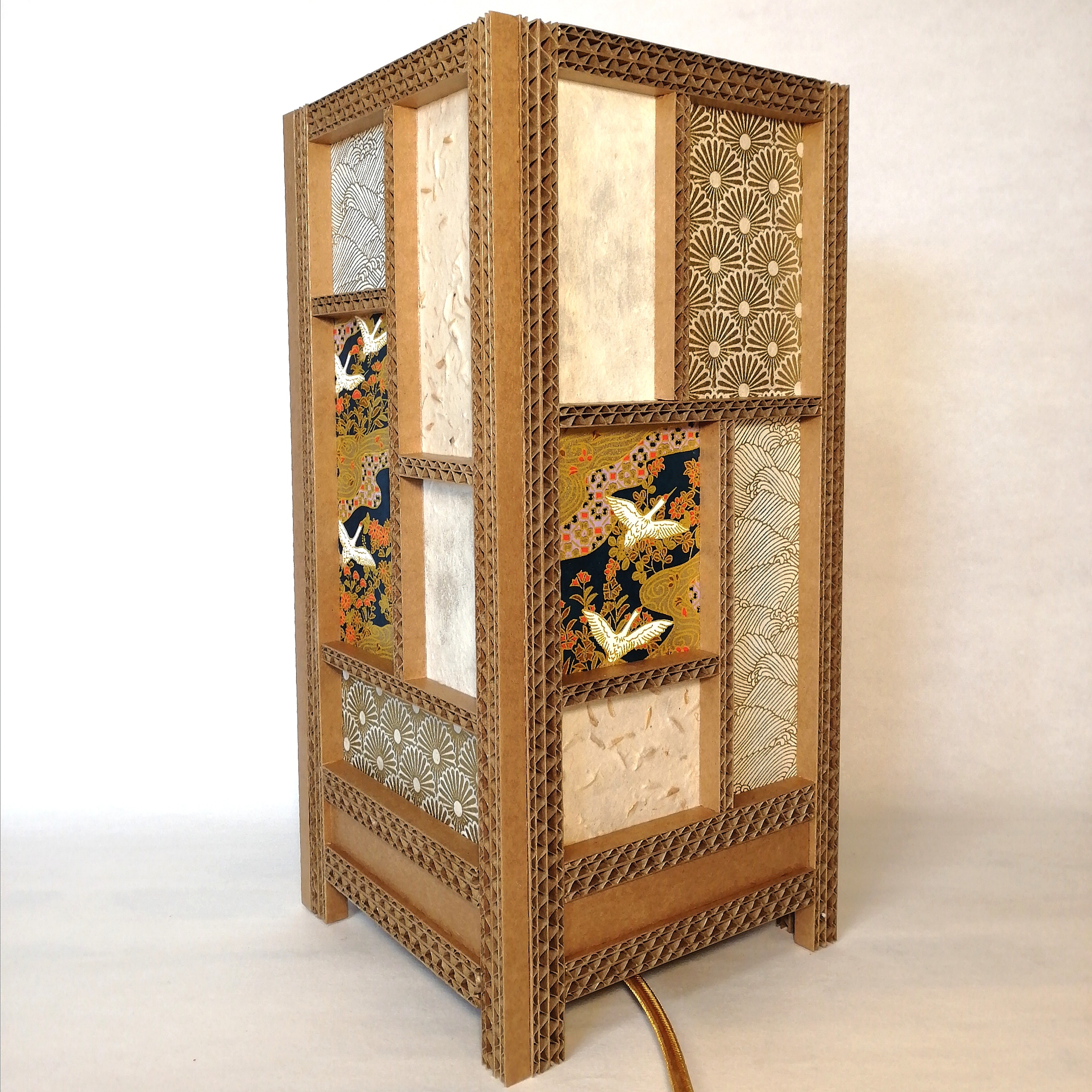 JEMA 2024-autour du carton- lampe grues :  lampe d'ambiance en carton, papier japonais et papier népalais. La structure de la lampe (carrée) est réalisée à partir de baguettes de carton ondulé sur lesquelles sont collés les différents papiers dans les tons crème et doré. Le principal motif sur cette lampe est la grue blanche qui s'envole.