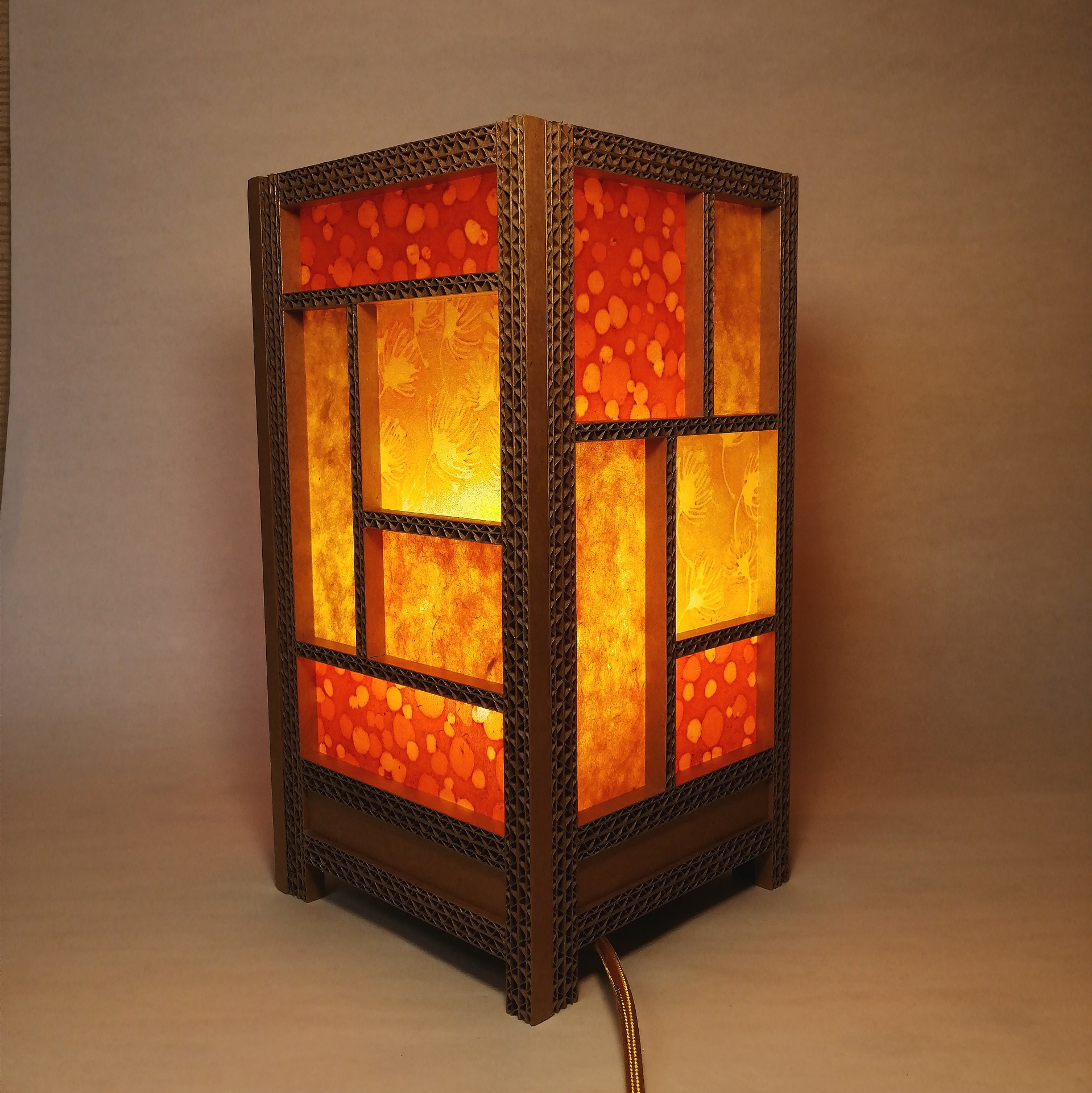 JEMA 2024-autour du carton-lampe orangée :  Lampe d'ambiance en carton et papier népalais et papier batik. La structure de la lampe (carrée) est réalisée à partir de baguettes de carton ondulé sur lesquelles sont collés les différents papiers dans les tons orangés.