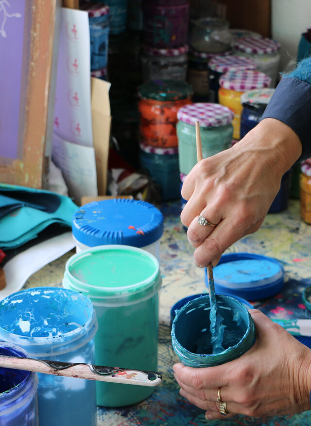 Marie B prépare des couleurs bleues dans son atelier de sérigraphie textile- détail de mains remuant un pot de couleur